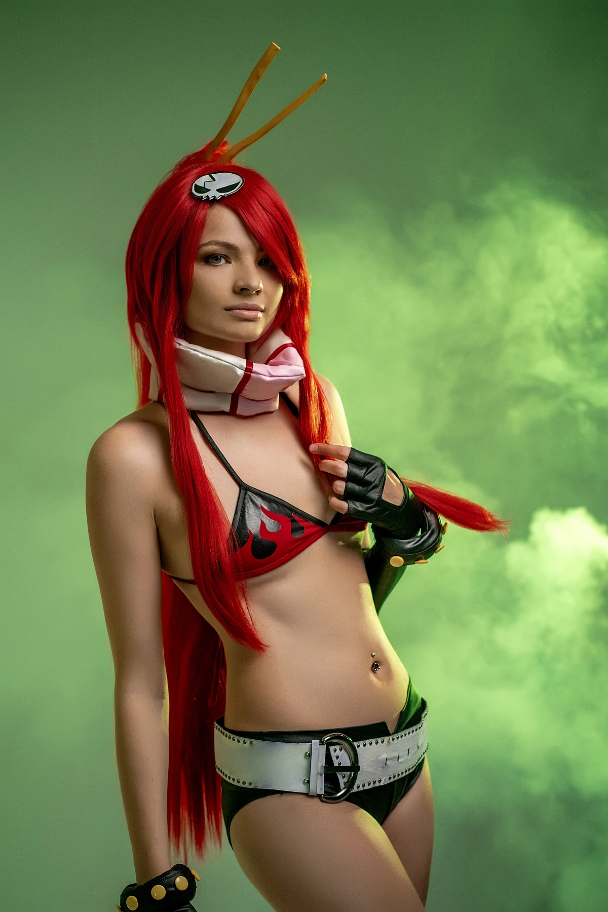 Red-haired cosplay girl in flame bikini touching hair amid green studio smoke background
